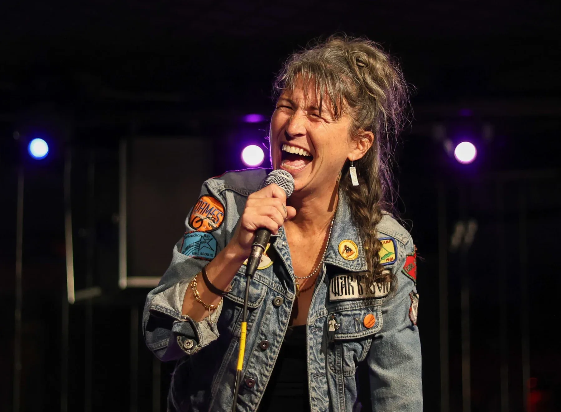 Pam Benjamin laughing onstage with a microphone in a patched denim jacket